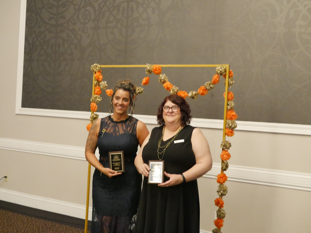 Photo of Jodi and Leslie. Jodi stands on the left wearing a dark grey lace gown and Leslie stands next to her in a black short sleeved dress. They stand in front of a gold arch draped with orange and gold paper flowers.